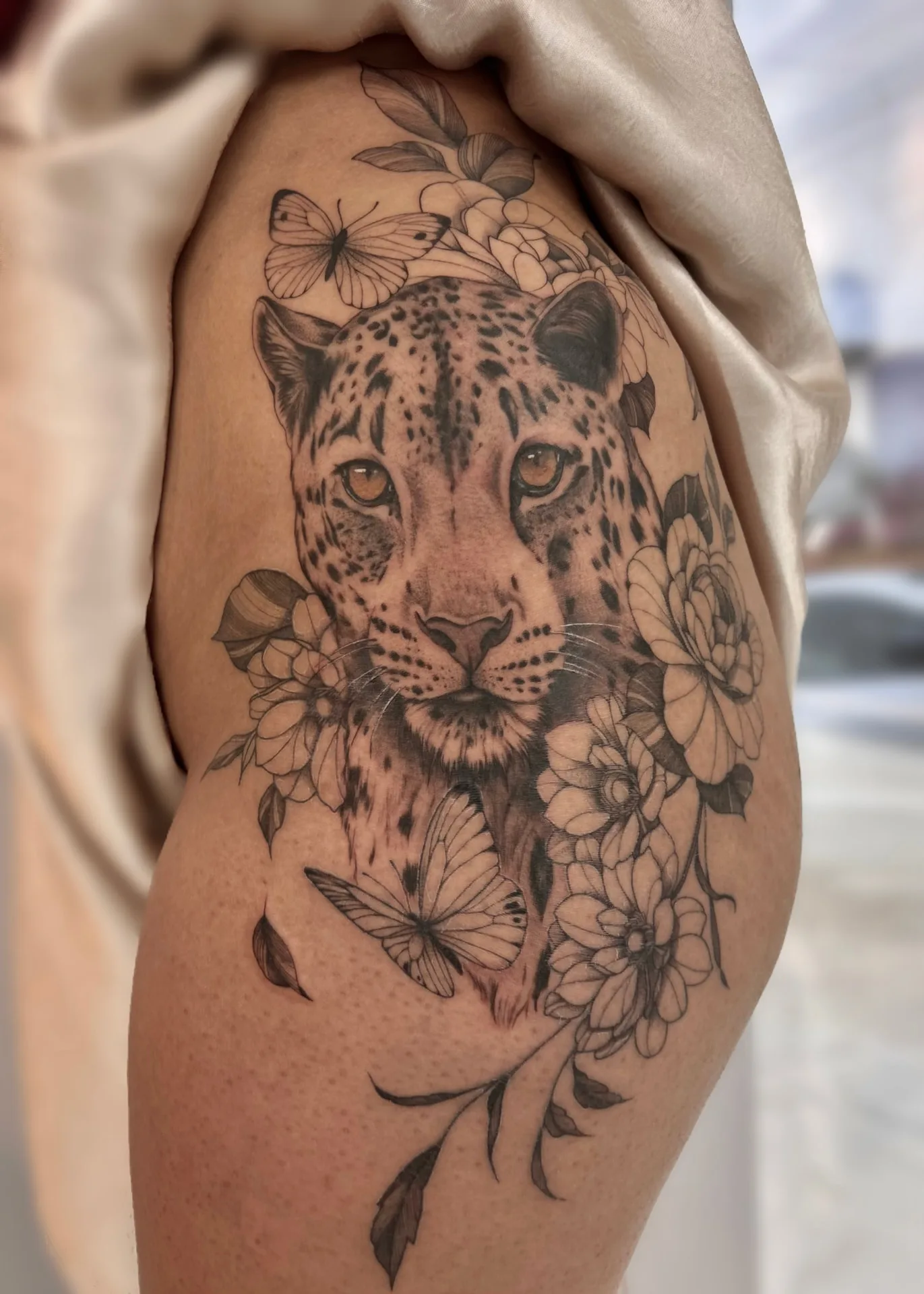 Tatuagem de onça com flores e borboletas em preto e cinza na coxa